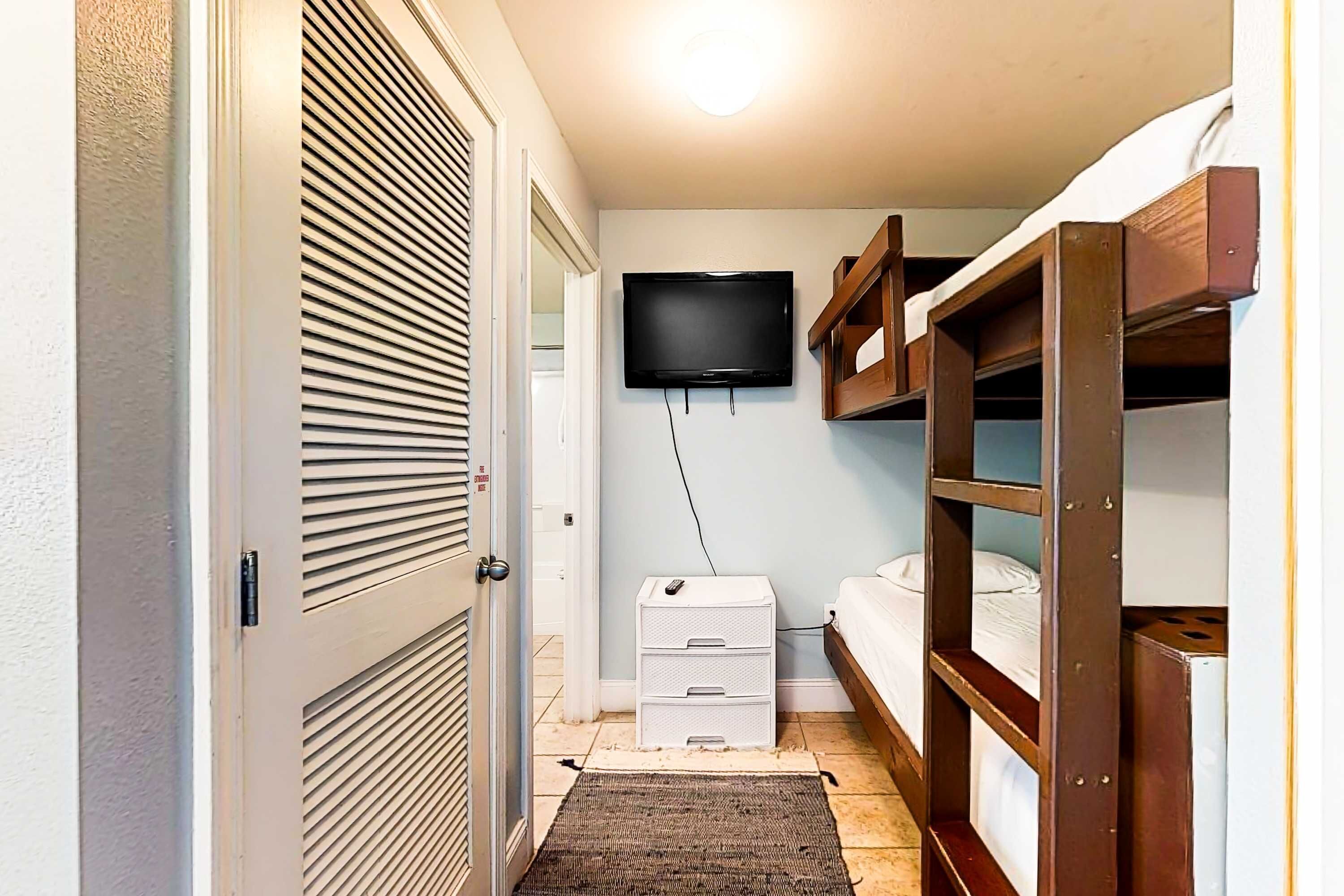 A bedroom with a bunk bed, a TV mounted on the wall, a small white dresser, and a door with a louvered design.