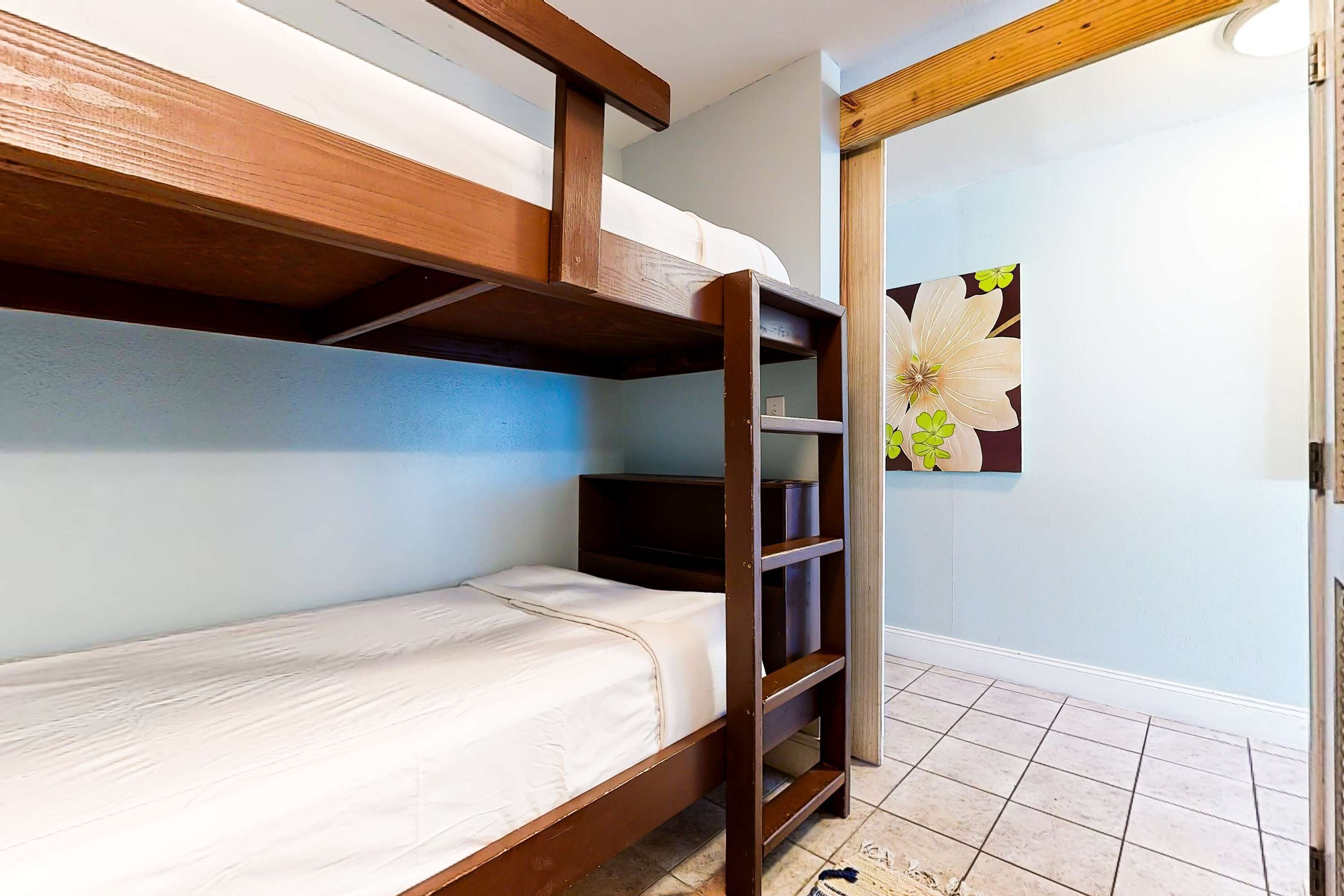 A bunk bed with a wooden frame and white bedding, a small shelf unit, and a door leading to another room.