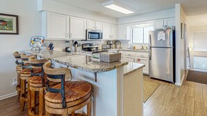 A kitchen with white cabinets, a stainless steel refrigerator, a microwave, and a coffee maker. There are wooden bar stools and a granite countertop. - Gorgeous Blue Mountain Beach Condo with views of the Gulf (Santa Rosa Beach)