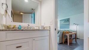 A bathroom with a marble countertop, a mirror, and a sink. There is a wooden table with a basket and a lamp in the adjacent room. - Gorgeous Blue Mountain Beach Condo with views of the Gulf (Santa Rosa Beach)