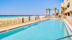 A swimming pool with a view of the ocean, palm trees, and a building with balconies. - Stunning beachfront condo with water views, pool, hot tub, & sauna (Panama City Beach)