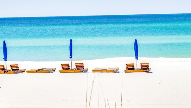 A beach with rows of wooden lounge chairs and blue umbrellas, arranged in a line facing the ocean. - Stunning beachfront condo with water views, pool, hot tub, & sauna (Panama City Beach)