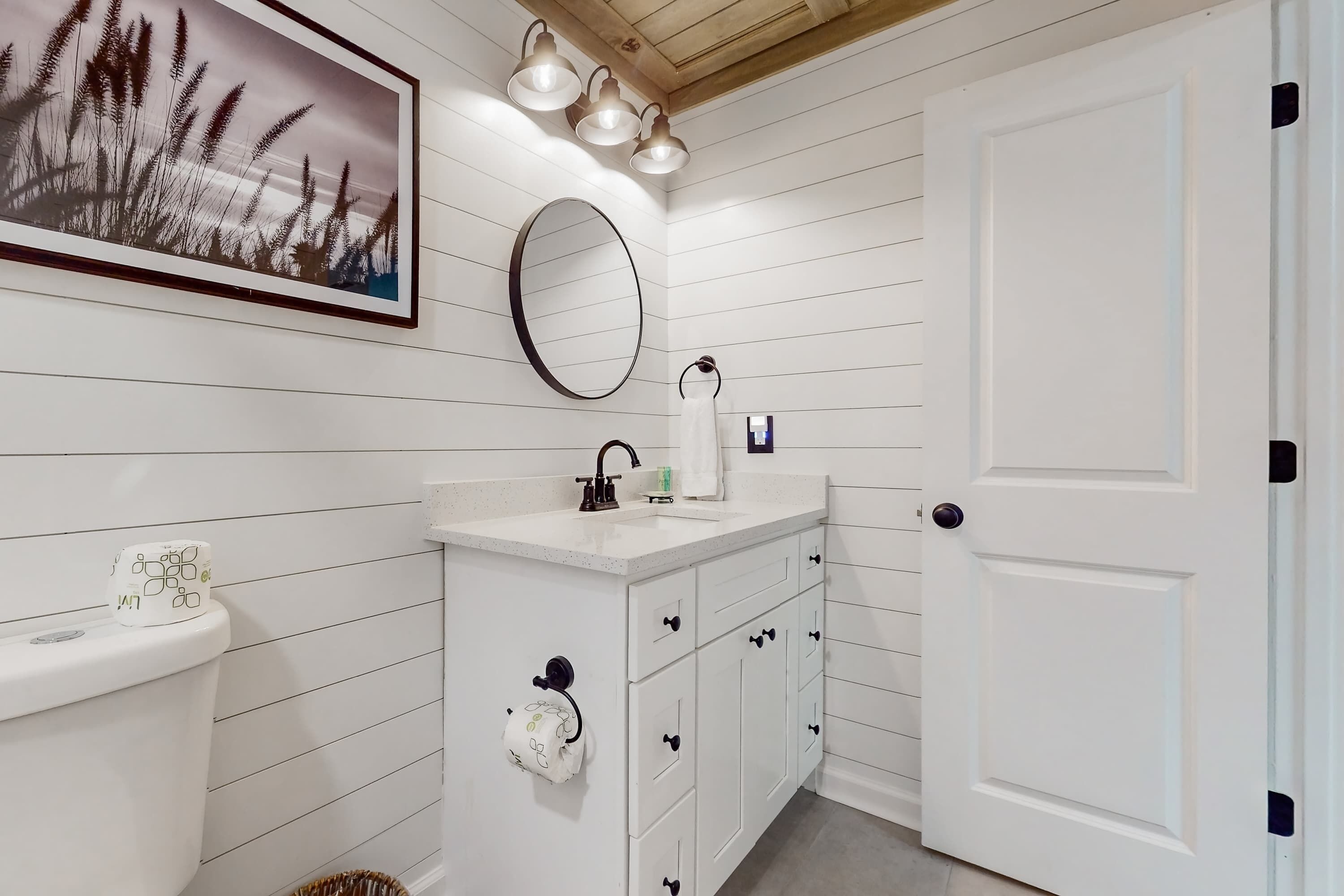 A bathroom with a toilet, sink, and mirror. There is a framed picture above the sink and a door to the right.