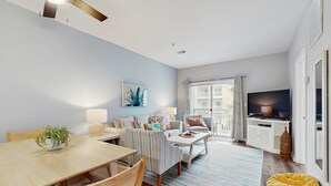 A modern living room with a dining area, a TV, and a large window with curtains. - Pool-view condo with private balcony, washer and dryer, Wifi, 2 bikes included (Santa Rosa Beach)