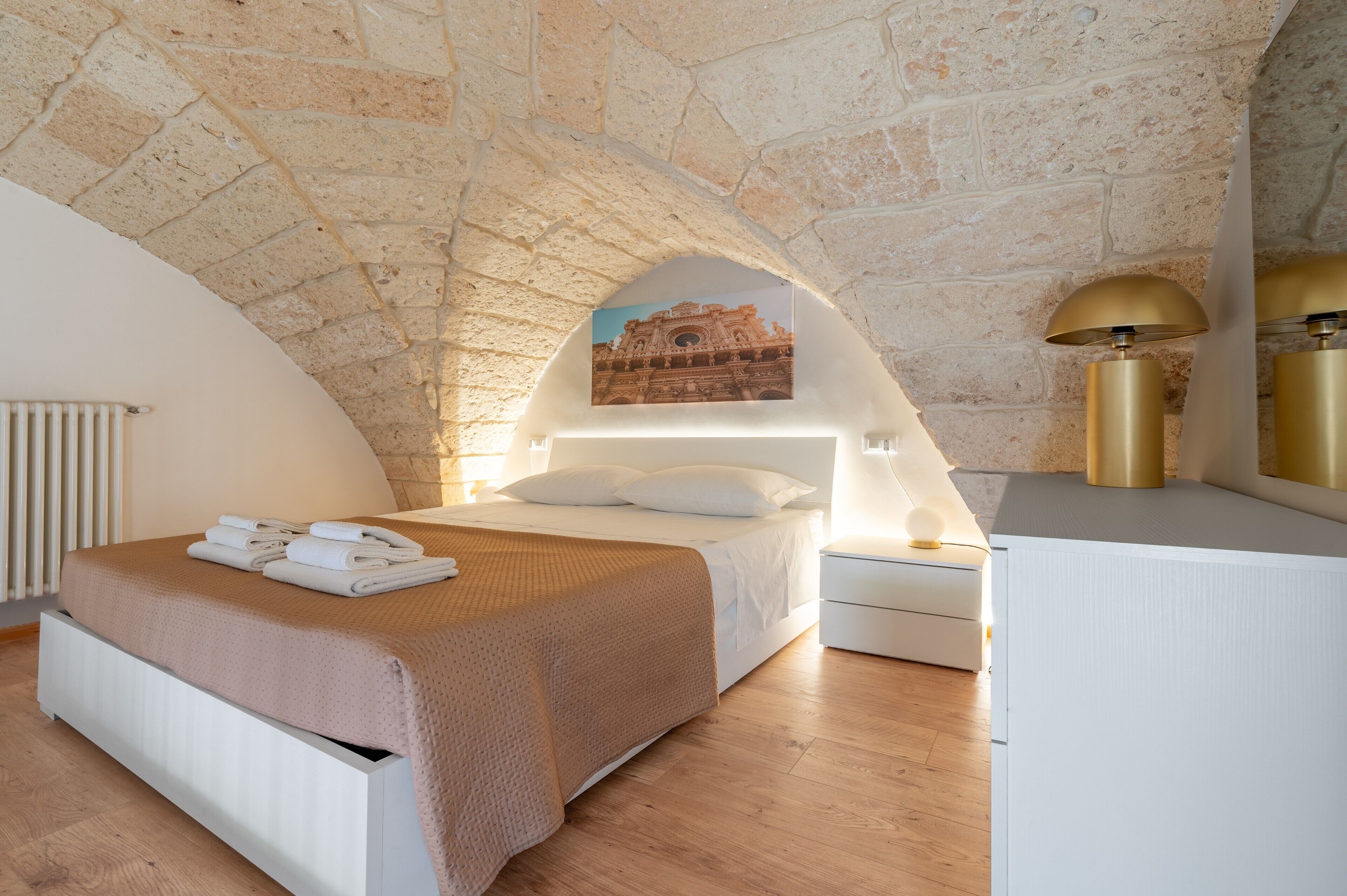 Ein Schlafzimmer mit einer Decke aus Steinbögen, einem Bett mit einer beige farbenen Bettdecke, einem Nachttisch mit einer Lampe und einem Wandbild.