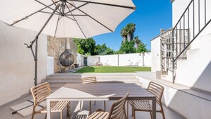 Een terras buiten met een tafel, stoelen, een grote parasol en een uitzicht op een tuin en gebouw op de achtergrond.