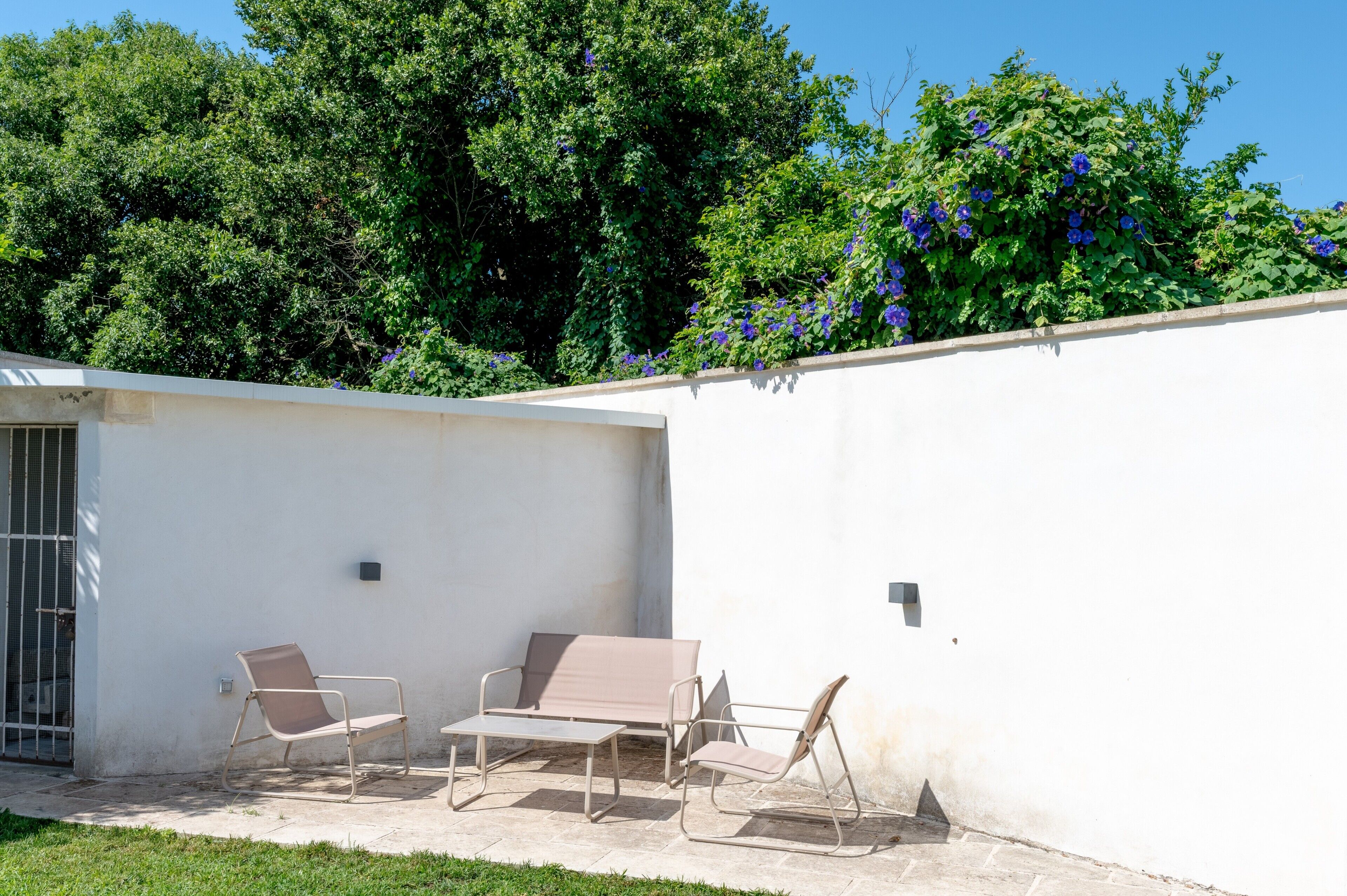 An outdoor patio with a white wall, a metal gate, and two beige lounge chairs with a small table.