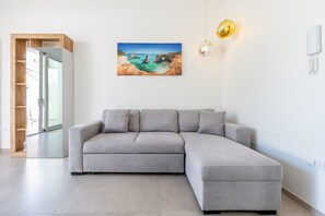 Un salon moderne avec un canapé d’angle gris, un tableau représentant un paysage côtier accroché au mur et une suspension décorative.