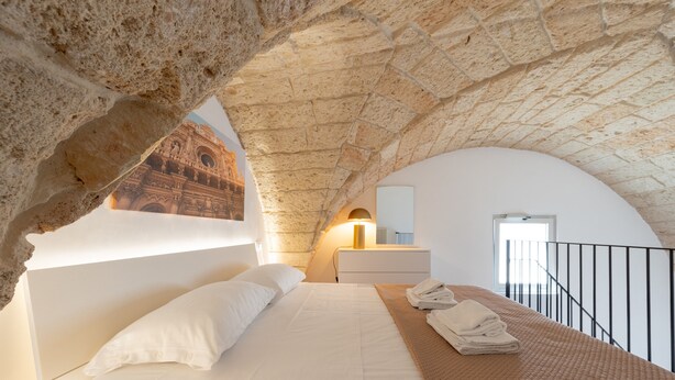 Een slaapkamer met een stenen plafond, een bed met witte lakens, een nachtkastje met een lamp, en uitzicht op een gebouw door een rondboogvenster.