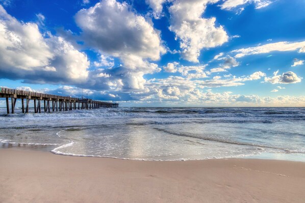Ein Strand mit einem hölzernen Steg, der sich ins Meer erstreckt, sanften Wellen, die an das Ufer schlagen, und einem teils bewölkten Himmel.