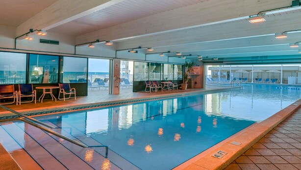 En indendørs swimmingpool med klart vand, omgivet af liggestole og borde, samt udsigt over byen gennem store vinduer.