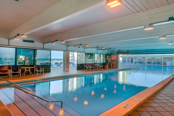 Ein Hallenbad mit klarem Wasser, umgeben von Liegestühlen und Tischen, mit Blick auf die Stadt durch große Fenster.