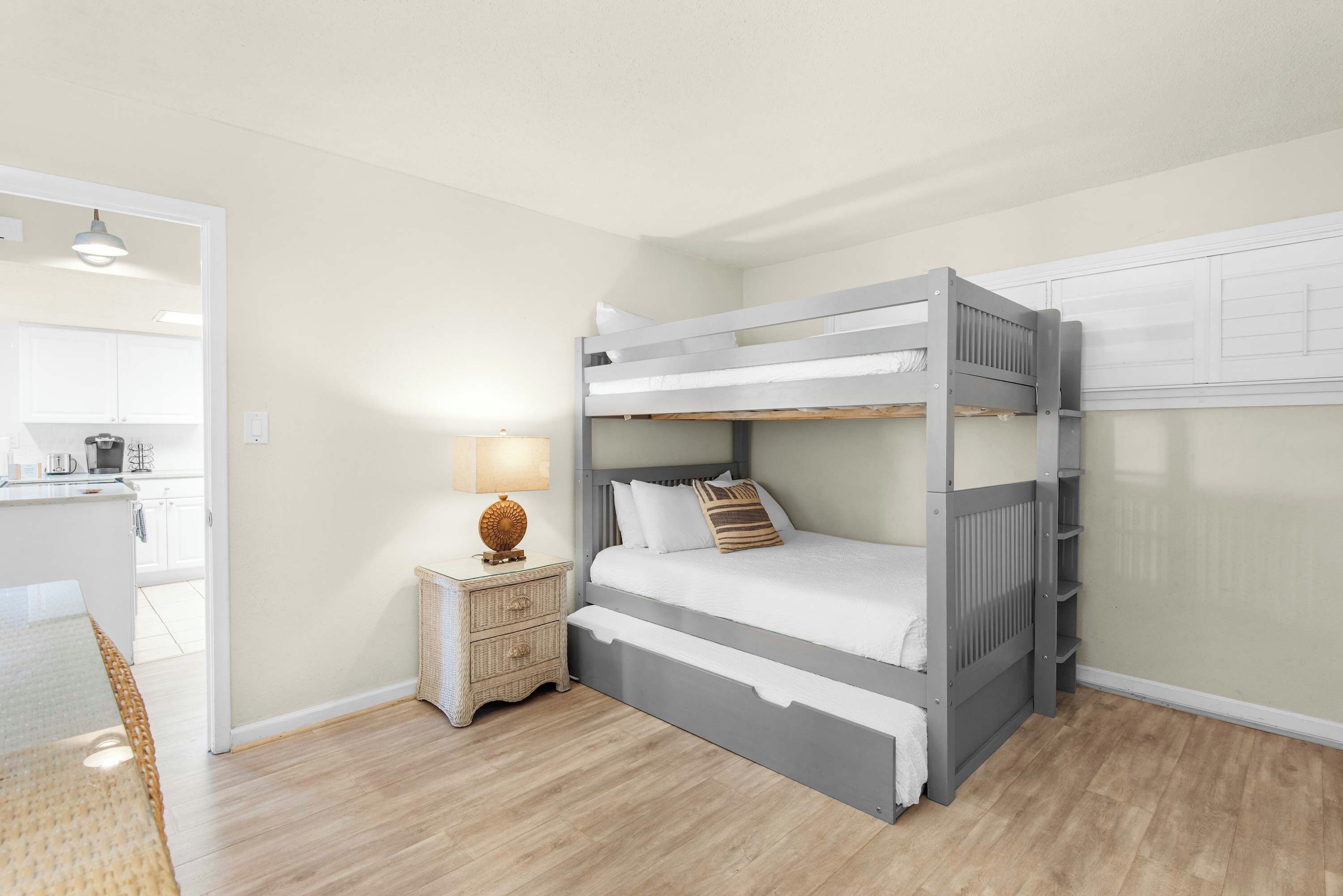 A bunk bed with a bedside table and lamp, a kitchen area with cabinets, and a wicker chair.