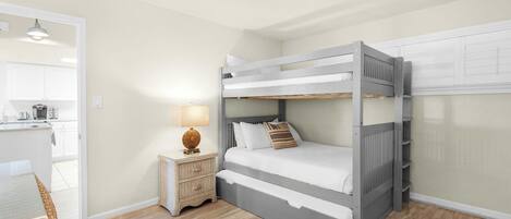 A bunk bed with a bedside table and lamp, a kitchen area with cabinets, and a wicker chair.