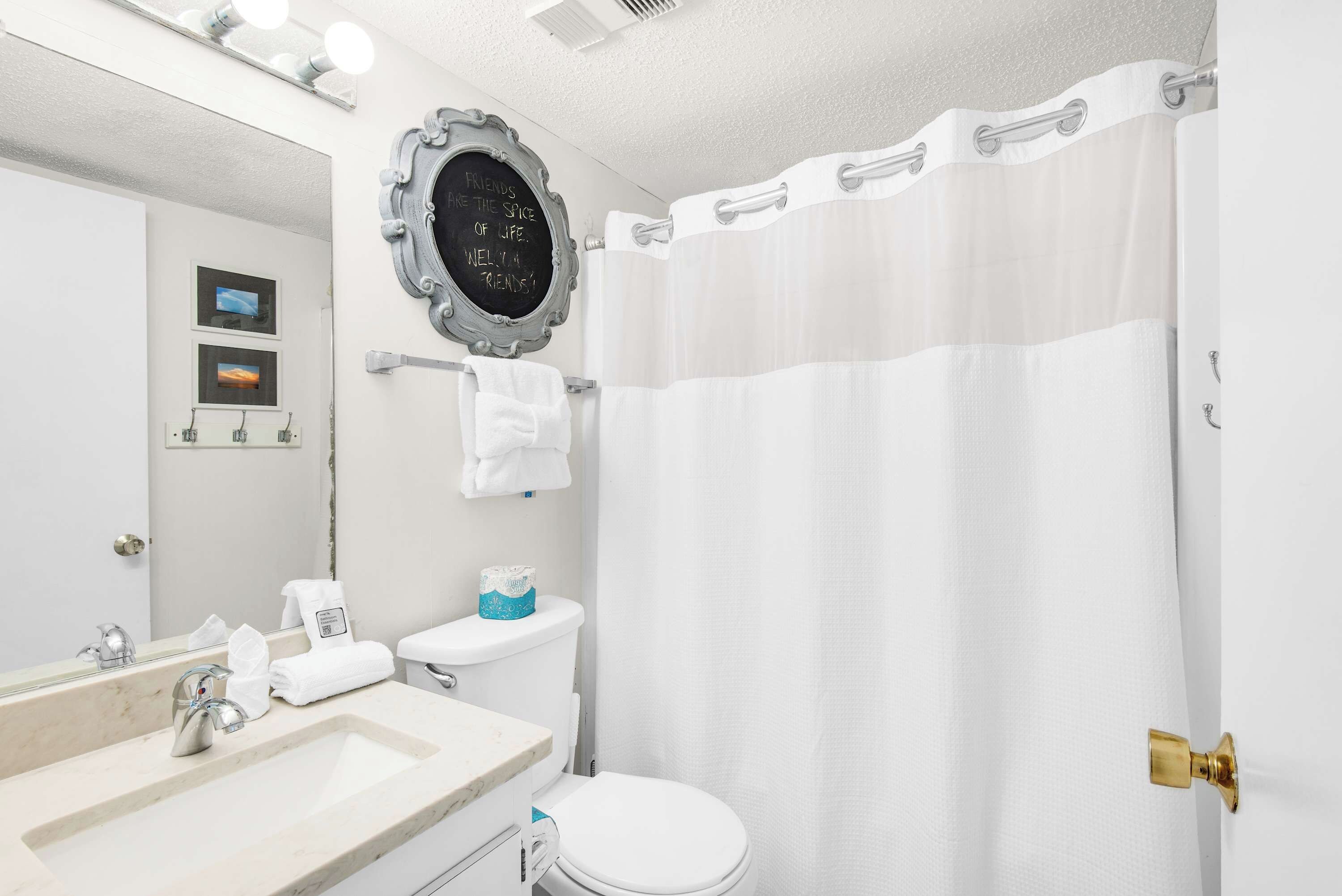 Une salle de bain avec un lavabo blanc, des toilettes, un rideau de douche et un miroir doté d’un cadre décoratif.
