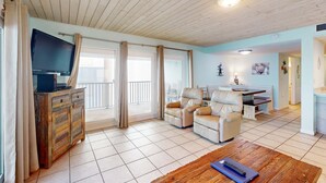 A spacious living area with a wooden ceiling, a television on a cabinet, a dining table with chairs, and a balcony with curtains. - Oceanfront condo with shared pool, resort amenities, and private washer/dryer (Panama City Beach)