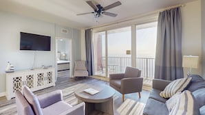 Una sala de estar moderna con una televisión de pantalla plana, un ventilador de techo, un sofá, sillones, una mesa de centro y un balcón con cortinas.