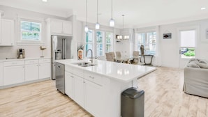 A modern kitchen with white cabinetry, a large island with a sink, and a dining area with a table and chairs. - Relaxing getaway outdoor pool access, gym, bikes, central AC, & W&D (Inlet Beach)