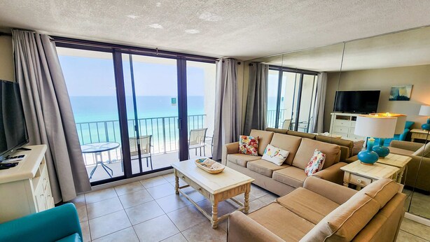 Uma sala de estar moderna com sofá, mesa de centro e televisão. O quarto tem grandes janelas com vista para o mar.
