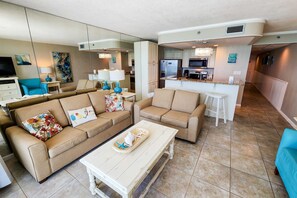 A modern living room with beige sofas, a coffee table, and a kitchen area with a refrigerator, microwave, and dining table. - Exquisite beachfront condo with pool, hot tub, balcony, central AC, & W/D (Panama City Beach)