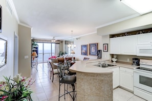 A modern kitchen with a central island, white cabinetry, and a dining area with chairs and a table. - Eclectic gulf front condo with free WiFI, two pools, hot tub, & easy beach access (Panama City Beach)