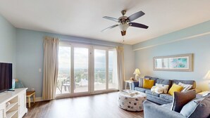A spacious living room with a large window, a ceiling fan, a grey sofa, and a small table with a lamp. - Beach condo with WiFi, amazing views, hot tub, and pools (Panama City Beach)