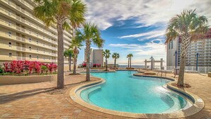 A hotel pool area with a curved pool, palm trees, and a building with balconies. - Beach condo with WiFi, amazing views, hot tub, and pools (Panama City Beach)