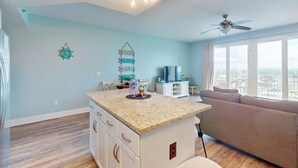 A modern living room with a kitchen island, a ceiling fan, and a large window offering a city view. - Beach condo with WiFi, amazing views, hot tub, and pools (Panama City Beach)
