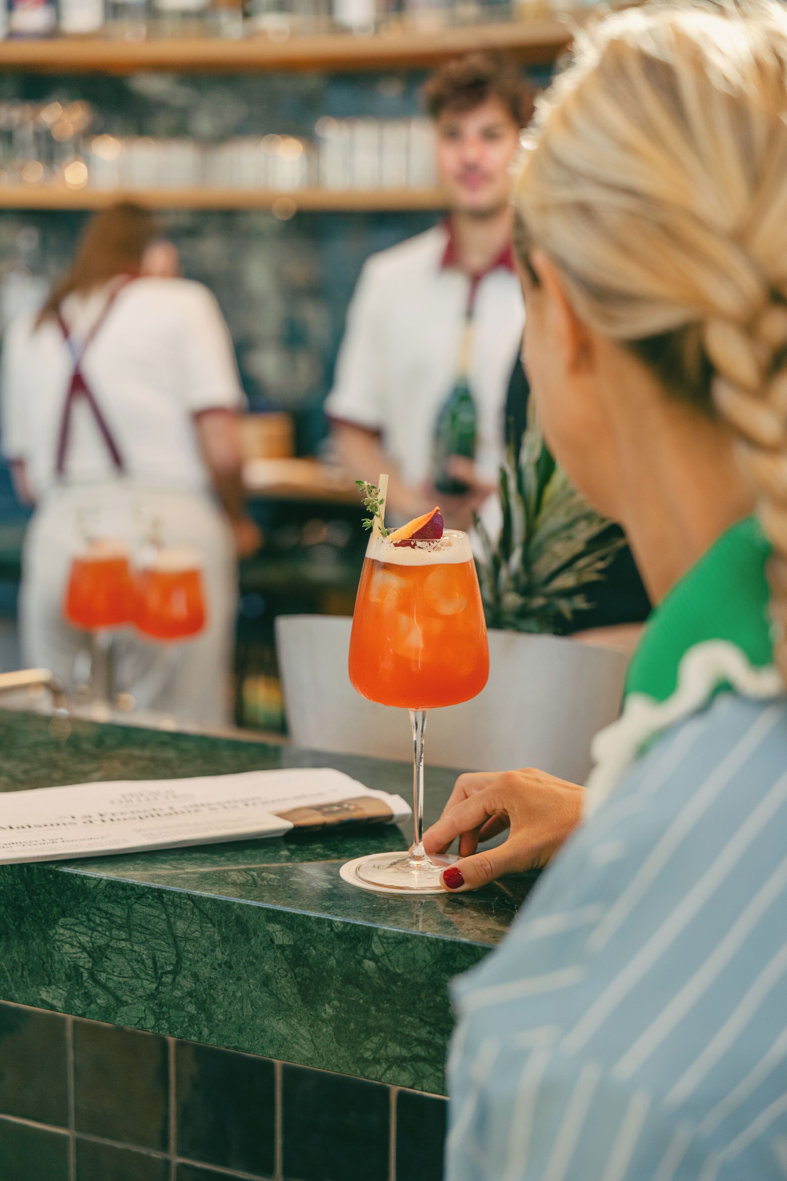 A bar area with a person holding a cocktail glass, a menu on the counter, and a blurred background with another person.