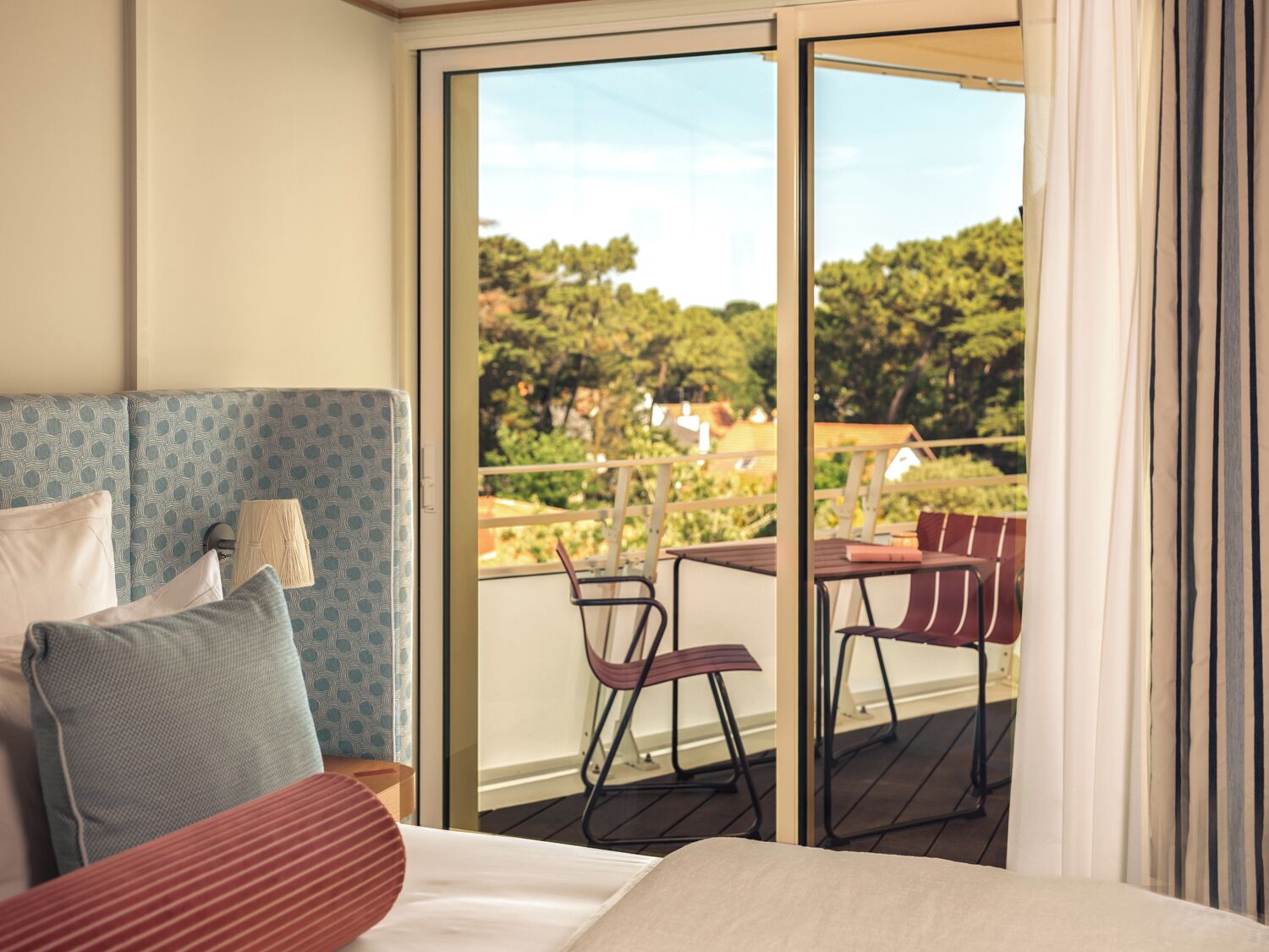 Habitación de hotel con cama, cabecera azul estampada, balcón con sillas y vistas a árboles y casas.