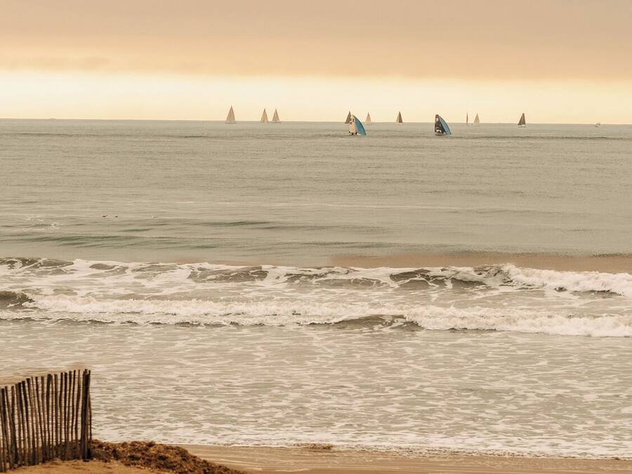 Una playa con una valla de madera, olas y veleros a lo lejos.
