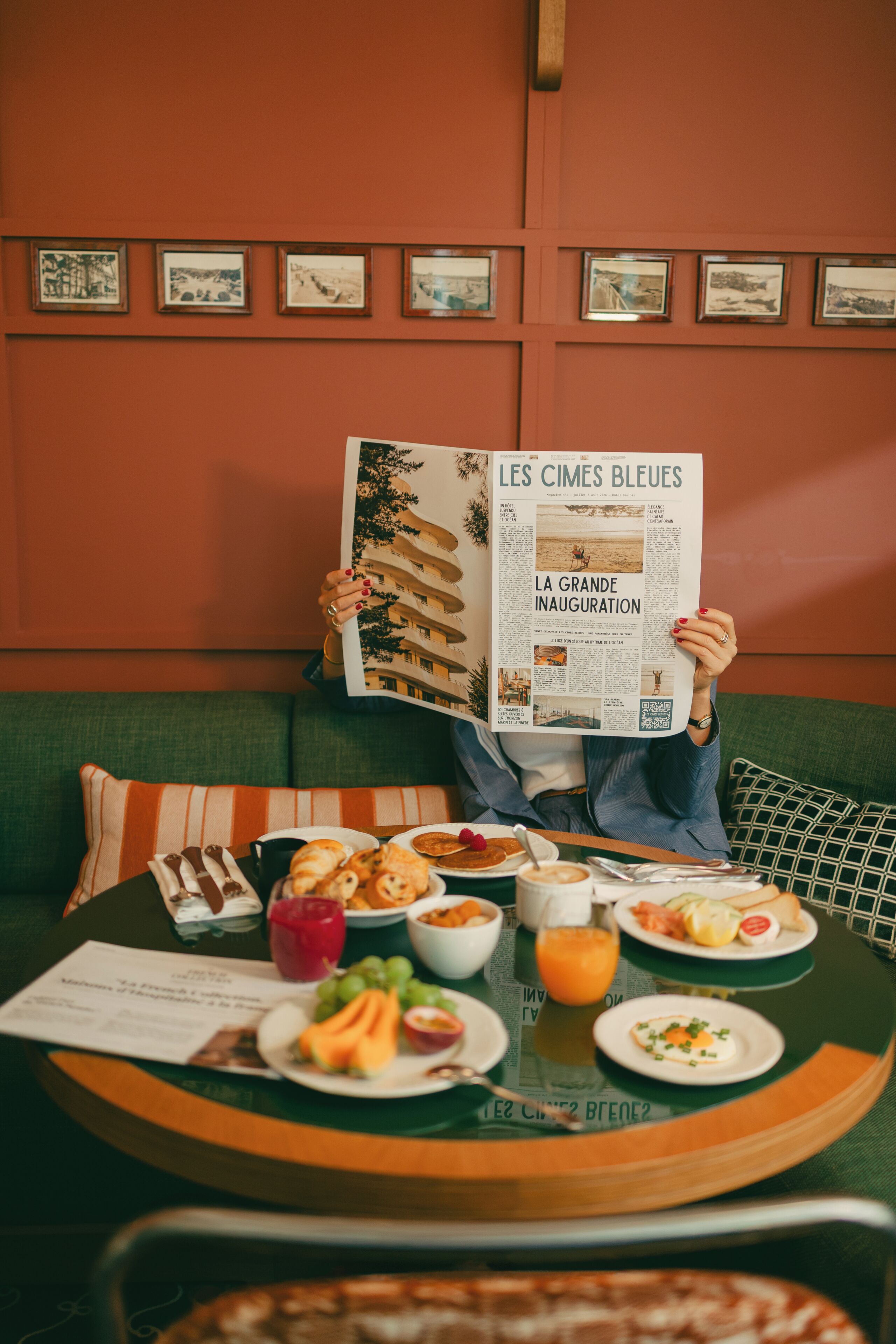 Une personne est assise sur un canapé vert et lit un journal. Une table se trouve devant elle, sur laquelle on peut voir un repas et un verre de jus d’orange.