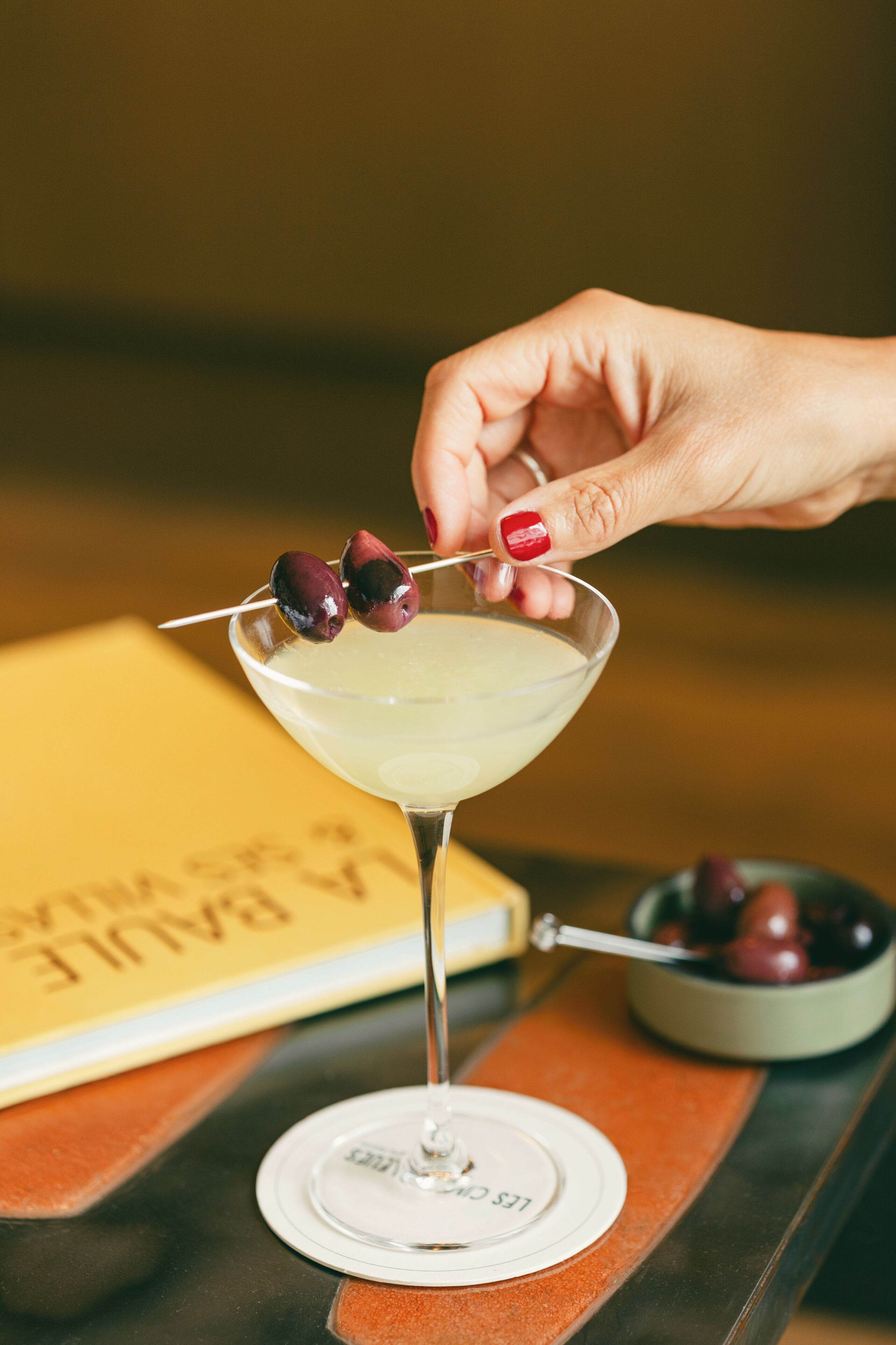A martini glass with olives on a cocktail stick, a book, and a bowl of olives on a table.