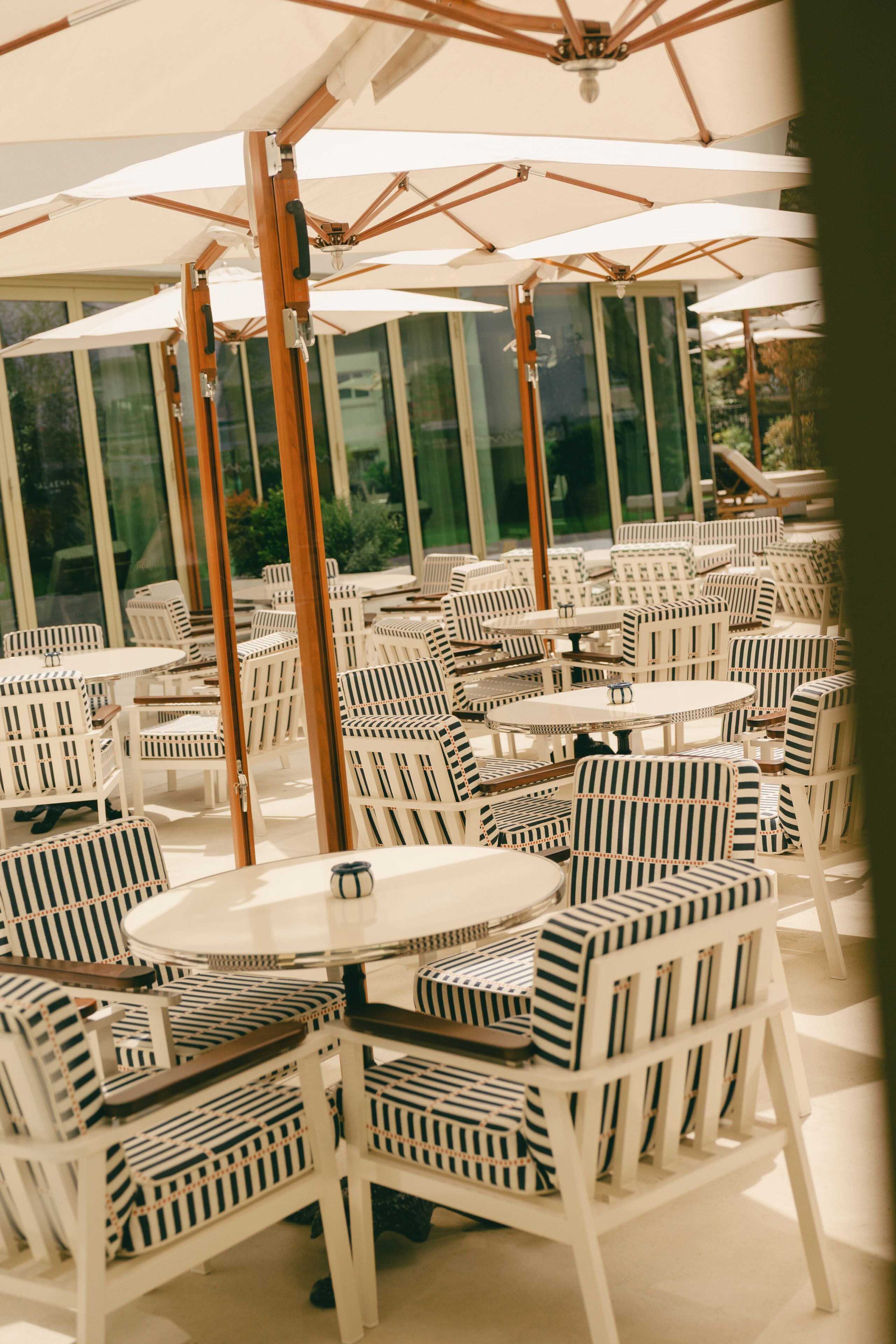 Un espace extérieur aménagé avec des chaises et des tables à rayures blanches, protégé par de grands parasols et entouré de baies vitrées.
