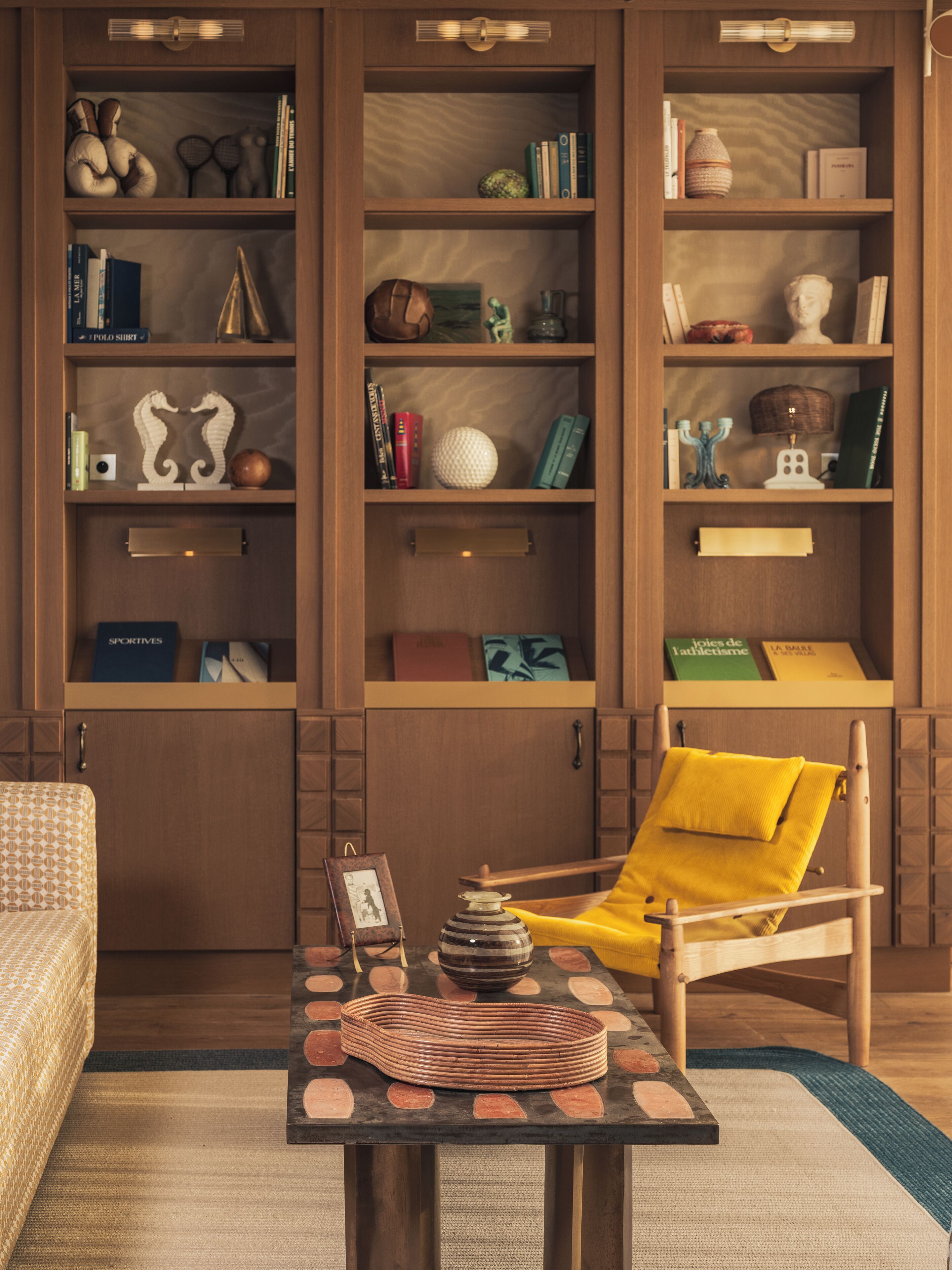 Een kamer met een houten boekenkast, een gele fauteuil, een klein tafeltje met decoratieve objecten en een kamerbreed tapijt met een patroon.