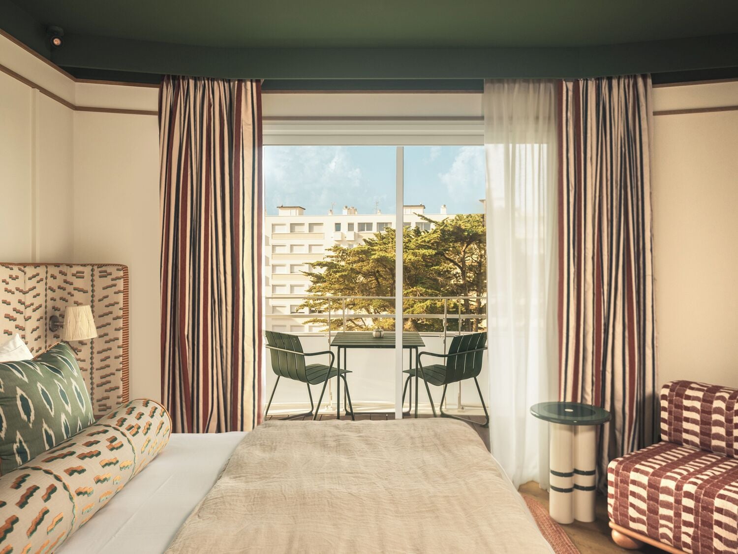 Habitación de hotel con una cama, dos sillas, una mesita y vistas a edificios y árboles.