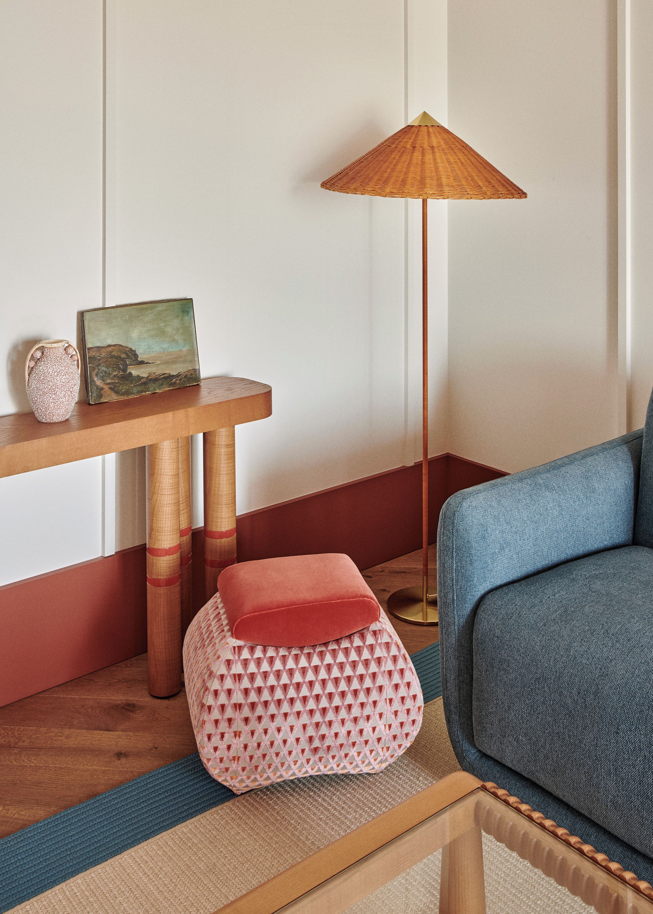 A room with a wooden side table, a floor lamp, a sofa, and a patterned ottoman.