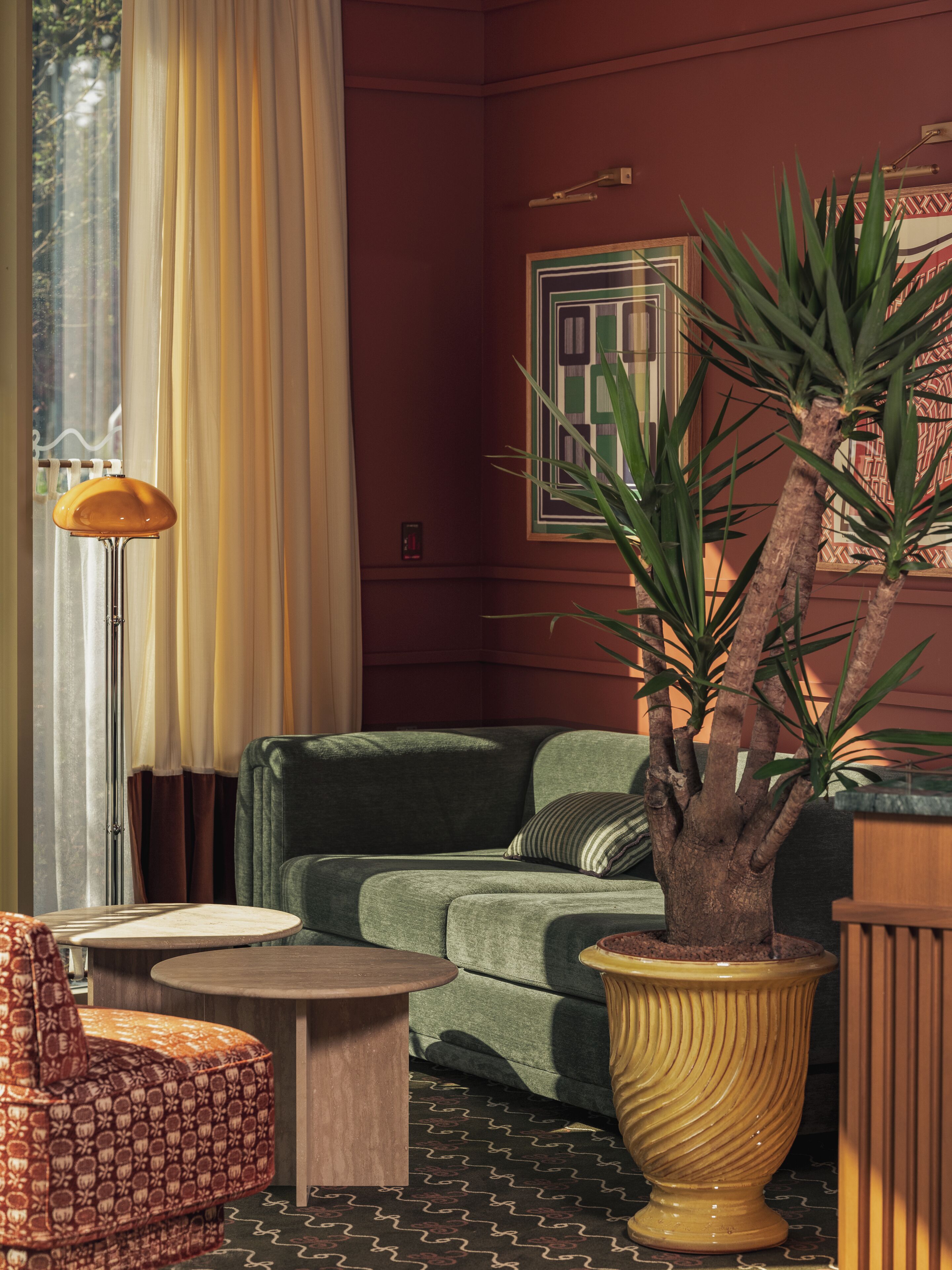 Une pièce avec un canapé vert, un fauteuil à motifs, une petite table ronde, un lampadaire et une grande plante en pot.