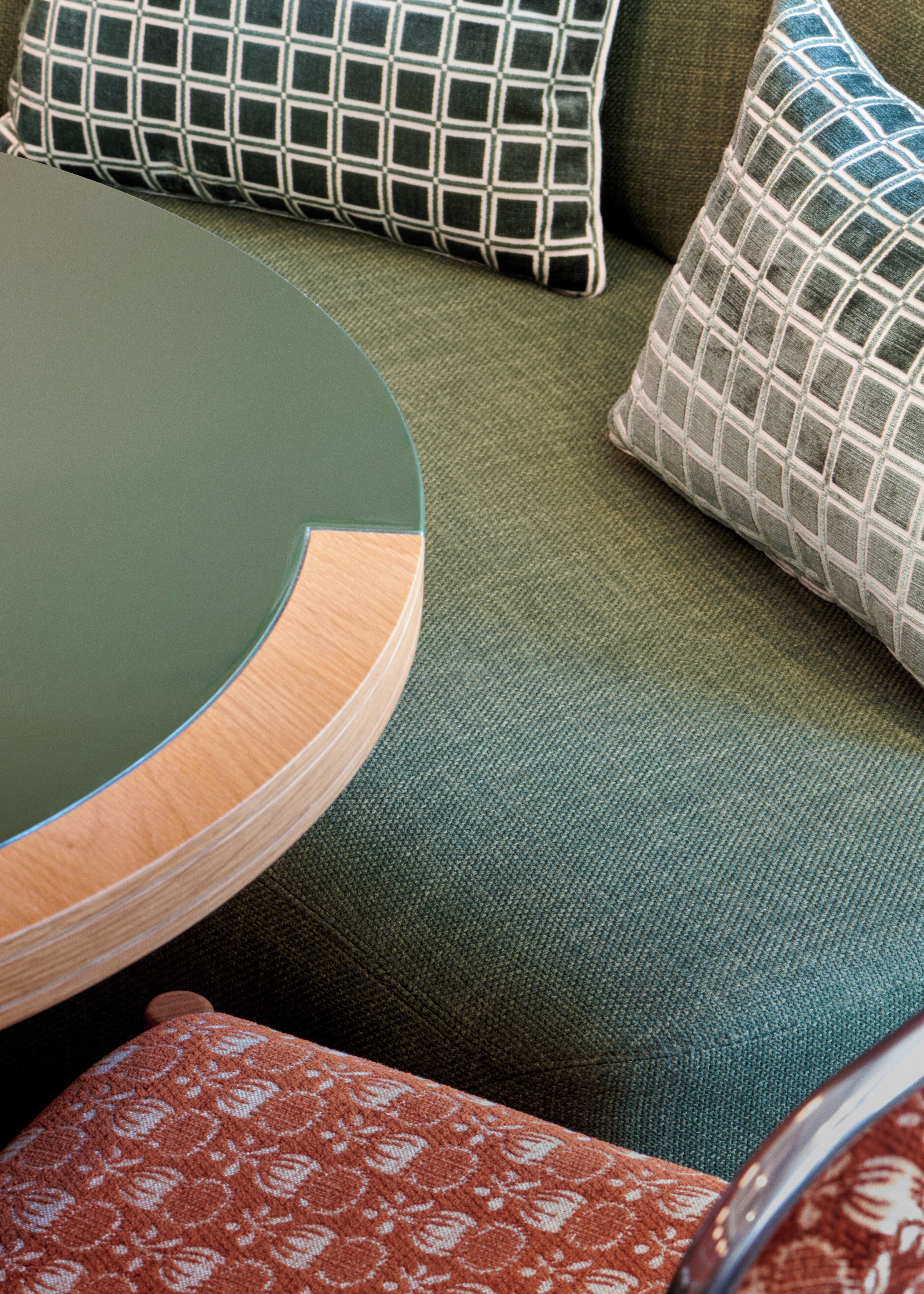 A round table with a green top and wooden base, a green sofa with patterned cushions, and a red patterned cushion.