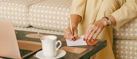 Une personne assise à une table, devant un ordinateur portable, en train d’écrire sur une feuille de papier, avec une tasse de café à portée de main.
