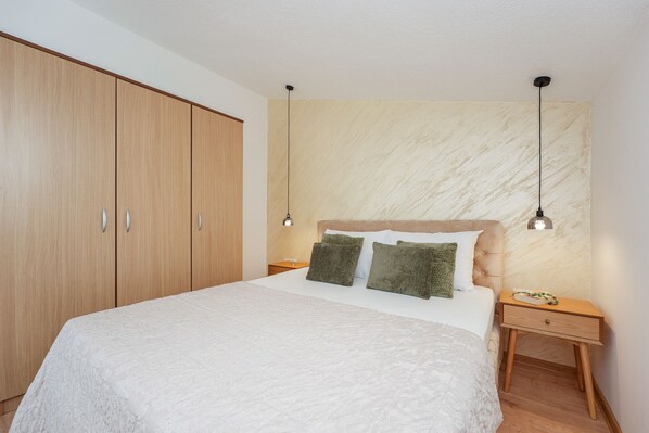 Une chambre à coucher avec un grand lit, une armoire en bois, deux tables de chevet avec des lampes et un mur décoré d’un motif imitant le marbre.