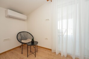 Une pièce avec un climatiseur fixé au plafond, une chaise noire avec un coussin, une petite table avec une plante et un rideau blanc transparent qui recouvre une fenêtre.