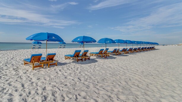 A beach with rows of wooden lounge chairs and blue umbrellas, arranged in a line along the shore. - Golf lovers’s condo with golf, 11 pools, hot tubs, gym, tennis, pickleball, W/D (Panama City Beach)
