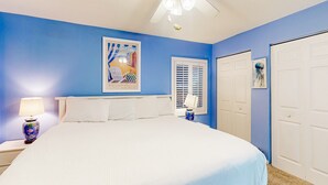 Une chambre à coucher avec un grand lit, un ventilateur de plafond, une table de chevet avec une lampe et une fenêtre avec des stores.
