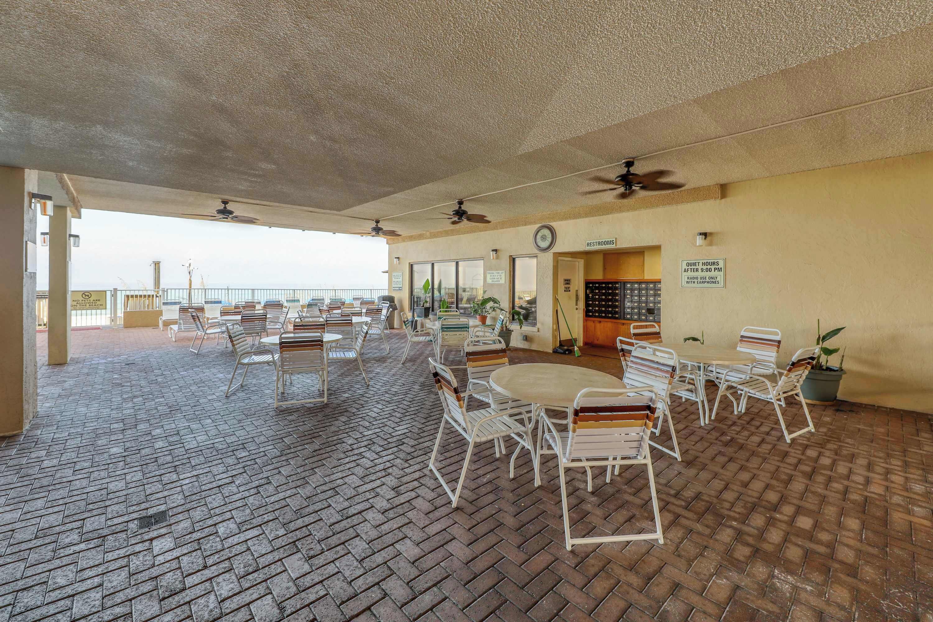 A covered outdoor seating area with brick flooring, white metal chairs and tables, ceiling fans, and a sign indicating guest hours.