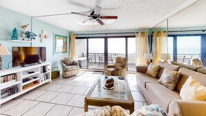 A spacious living room with a large flat-screen TV, comfortable seating, a glass coffee table, and a balcony with a view. - Coastal Gem w/ Beach & Pool Access, Oceanfront Views from Balcony, and Netflix (Panama City Beach)