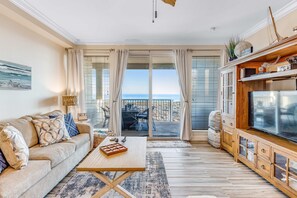 A living room with a sofa, coffee table, TV, wooden cabinet, and a balcony with a view of the sea. - Blissful Coastal Condo with Shared Pool, Ocean View, Easy Beach Access & WiFi (Navarre Beach)
