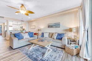 Ein modernes Wohnzimmer mit zwei Sofas, einem Couchtisch, einem Deckenventilator, einer Küche im Hintergrund und einem Korbwäscheschrank mit Handtüchern.
