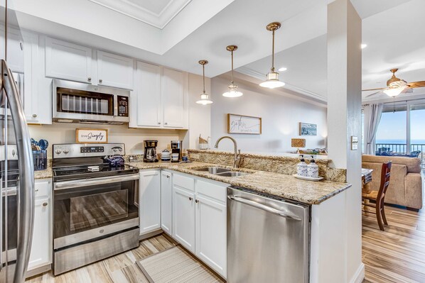 A modern kitchen with white cabinets, stainless steel appliances, and a granite countertop. There is a dining area with a view of the ocean. - Blissful Coastal Condo with Shared Pool, Ocean View, Easy Beach Access & WiFi (Navarre Beach)