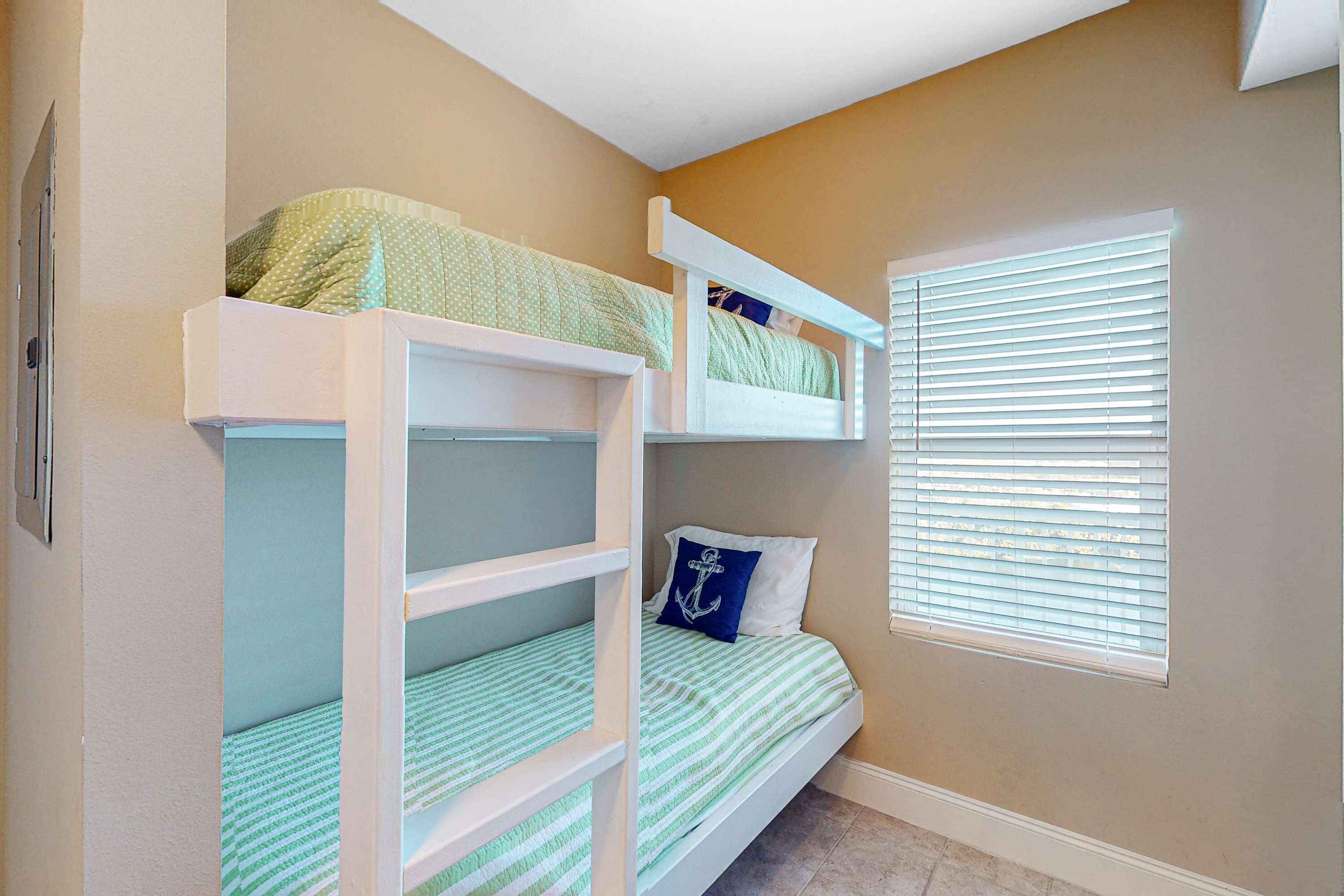 A room with bunk beds, a window with blinds, and a wall-mounted light switch.